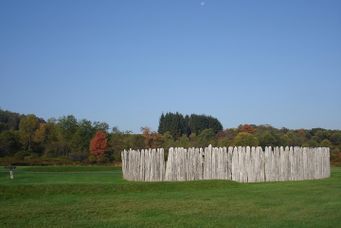A round fort made of sticks of wood rises from a green meadow with woods in the background and a clear blue sky.