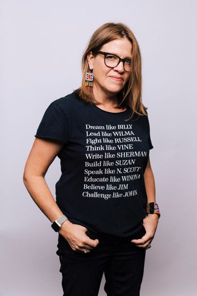 Maria Watt stands with her thumbs in her pockets. She wears black pants and a black t-shirt, black plastic rimmed glasses and native american earrings.