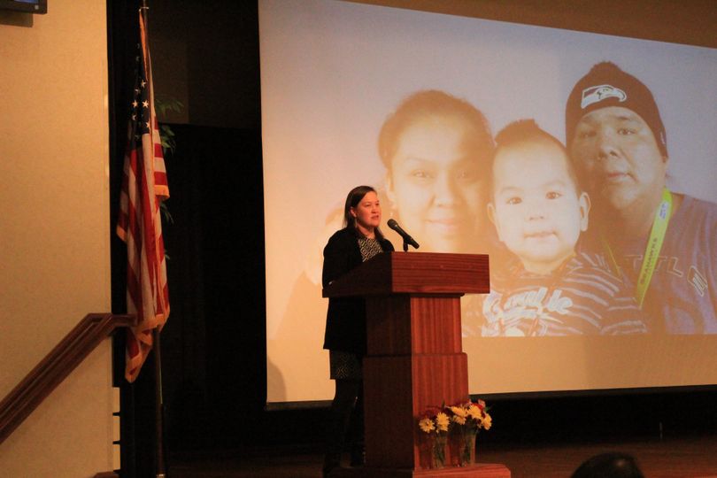 Speaker during the 2018 ceremony stands behind a podium and in front of a large projection of an indigenous family with father, mother and baby. 