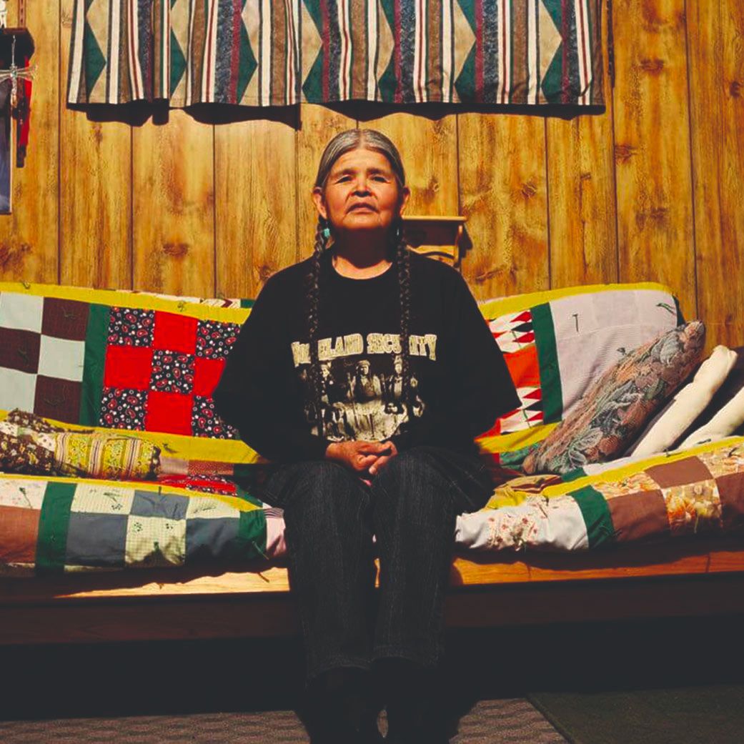 Jean Whitehorse sits on a futon with brightly patterned blanket. She wears all black and has grey hair styled in two braids. 
