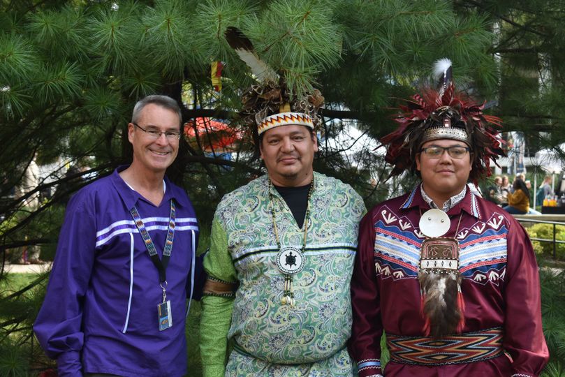 Three indigenous men stand together. The first man has light grey hair, wire glasses and wears a bright purple shirt and pants with a patterned stripe. The other two men wear a headband with feathers and patterned shirts
