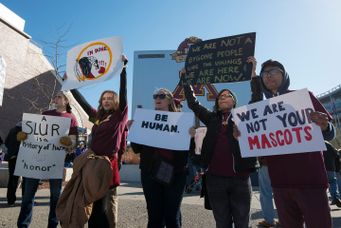 A group of five individuals stand outside of a football stadium holding signs to speak out against mascots using native american imagery.