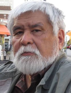 Male with bright white hair, mustache and beard stares intently into the camera. 