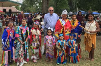 Man in a button down blue shirt, white hair and darkened glasses stands with a group of children dressed in their tribal regalia